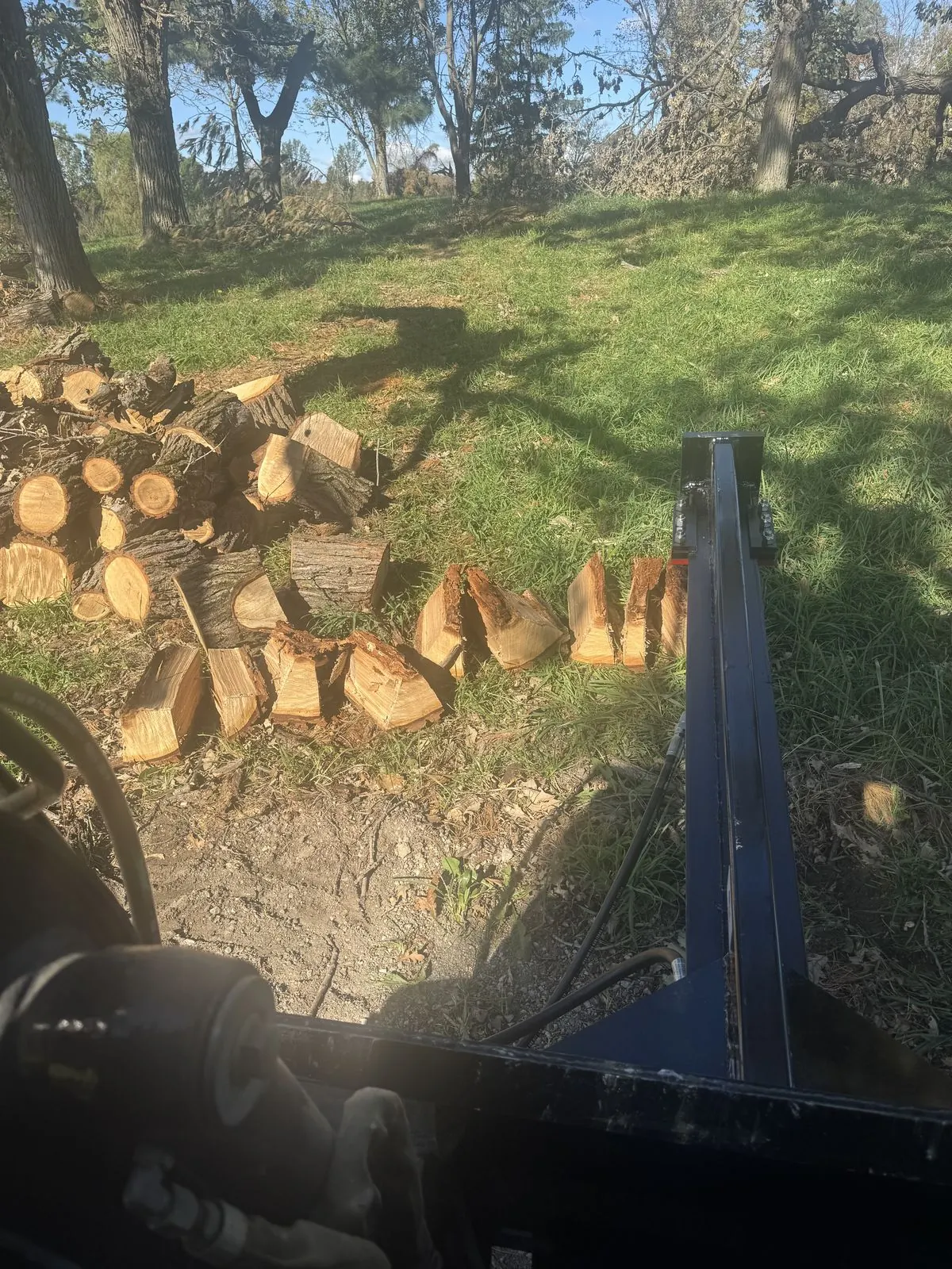 Split firewood on the grass, seen from the cab of the skid steer with its log splitter attachment